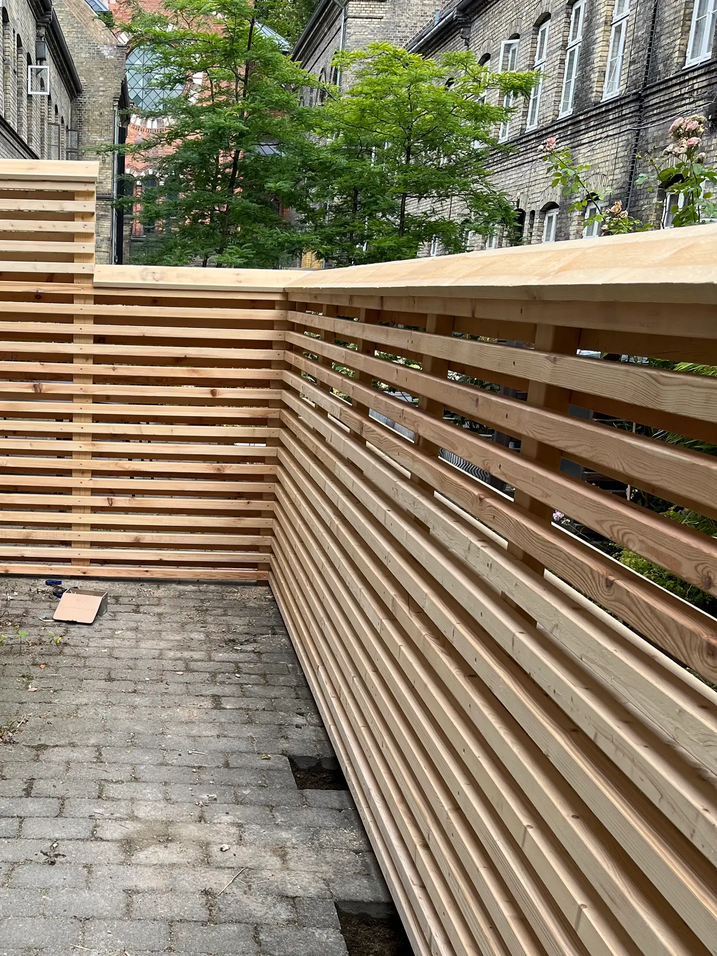 Lamelhegn i lyst, ubehandlet træ ført præcist rundt om hjørnet i en københavnsk gård med gulstensfacader og gråt fliseareal