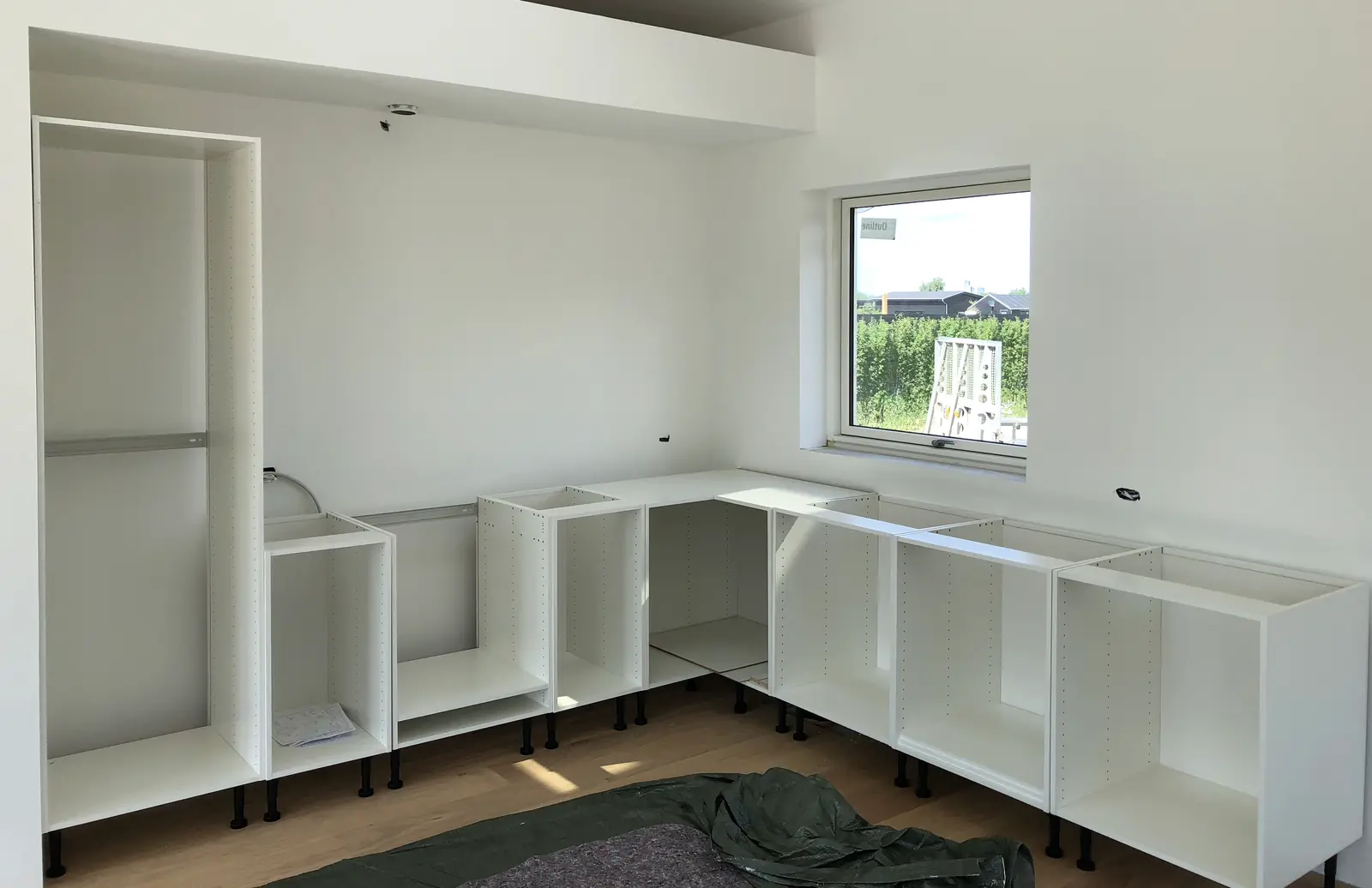 Køkkenet under montering med hvide korpus opstillet på justerbare ben i L-form, uden fronter og uden bordplade, egegulv
