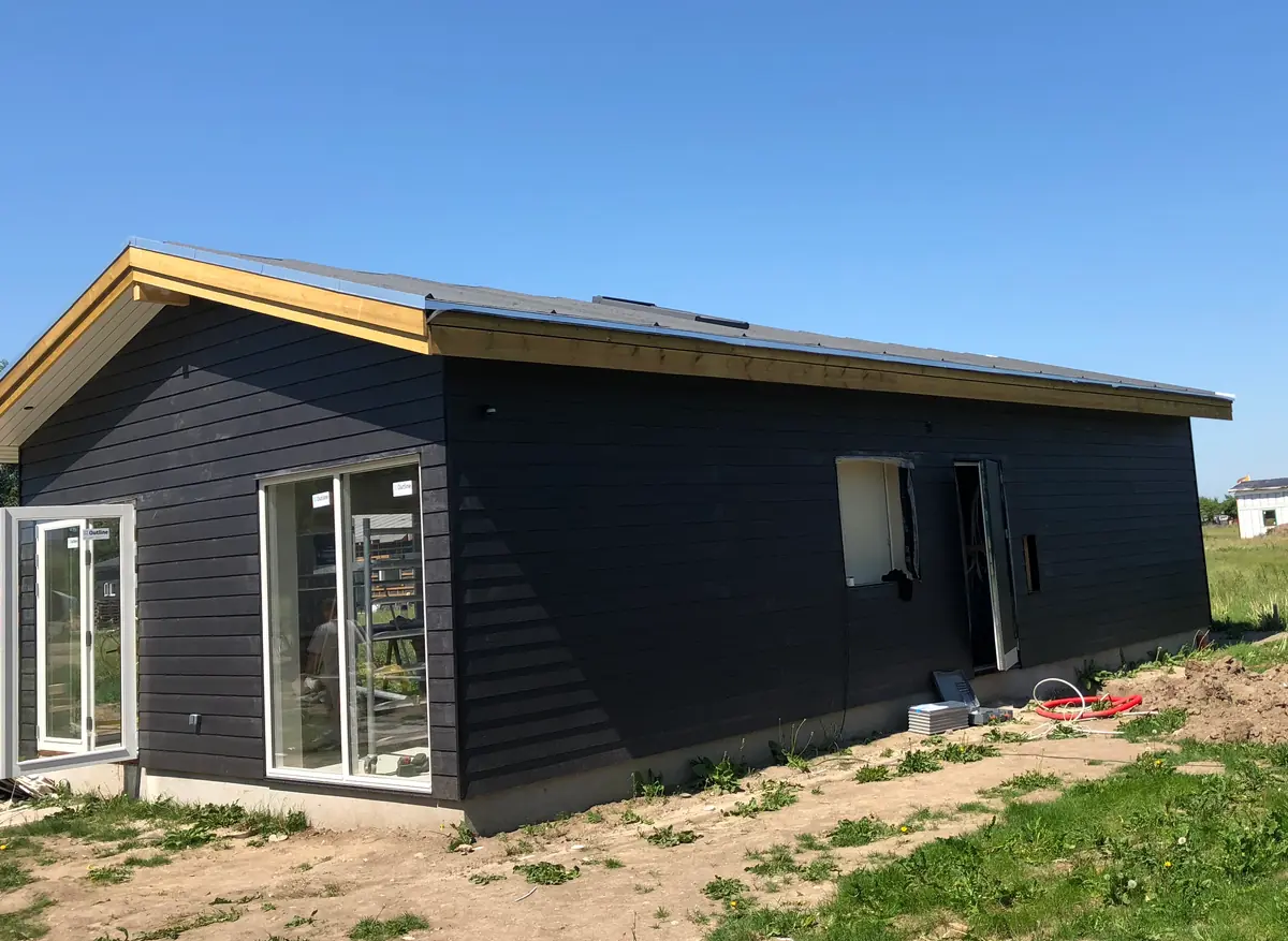 Hus med sort vandret træbeklædning, lyse spærender, lavt tag og hvide terrassedøre på sandet grund