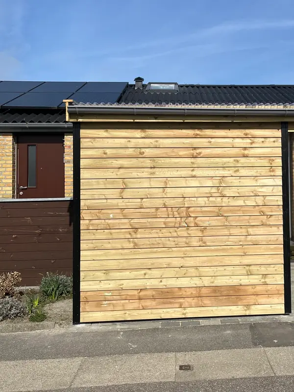 Den lukkede del af bygningen set forfra fra fortovet, med ubehandlet vandret klinkbeklædning mellem sorte hjørnelister, brunt hegn og rødbrun dør i gulstensfacaden til venstre