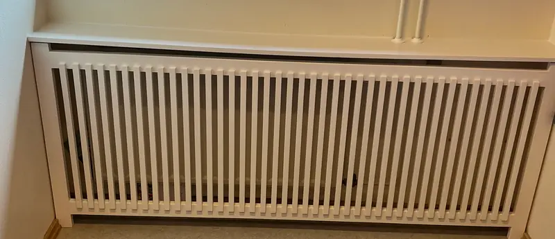 Hvid radiatorskjuler med lodrette tremmer og gennemgående hyldeplade, bygget ind mod væggen