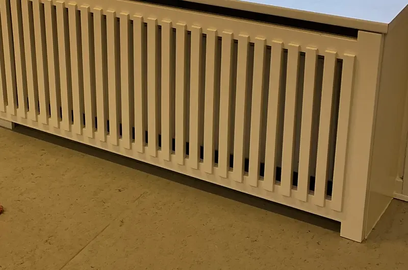 Hvid radiatorskjuler med lodrette tremmer og bred hyldeplade, set på skrå langs sin fulde længde
