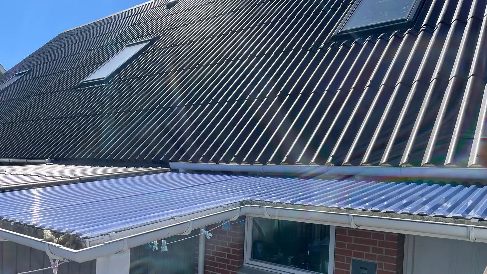 Sort bølgepladetag med to ovenlysvinduer og hvid tagrende over et gårdrum med klare tagplader og rød murstensfacade