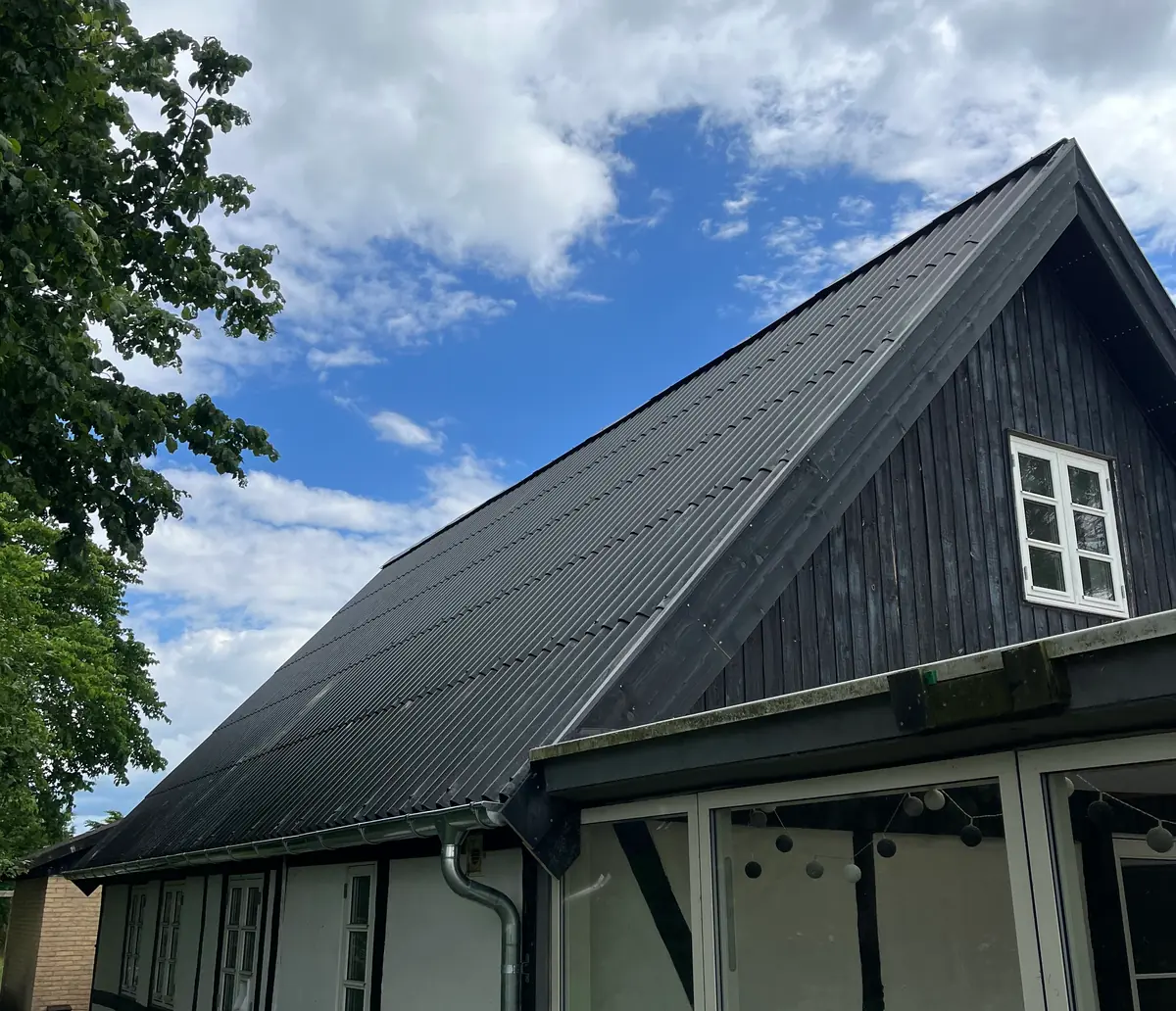 Sortmalet bræddegavl med nyt mørkt bølgepladetag, hvidt sprossevindue og zinktagrende, ved siden af hvidt bindingsværk