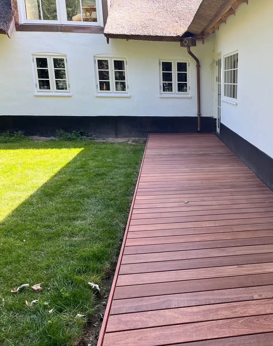 Terrasse i mørkt, rødbrunt træ ført langs den hvide facade på et stråtækt hus, med hvide sprossevinduer og kobbernedløb over dækket