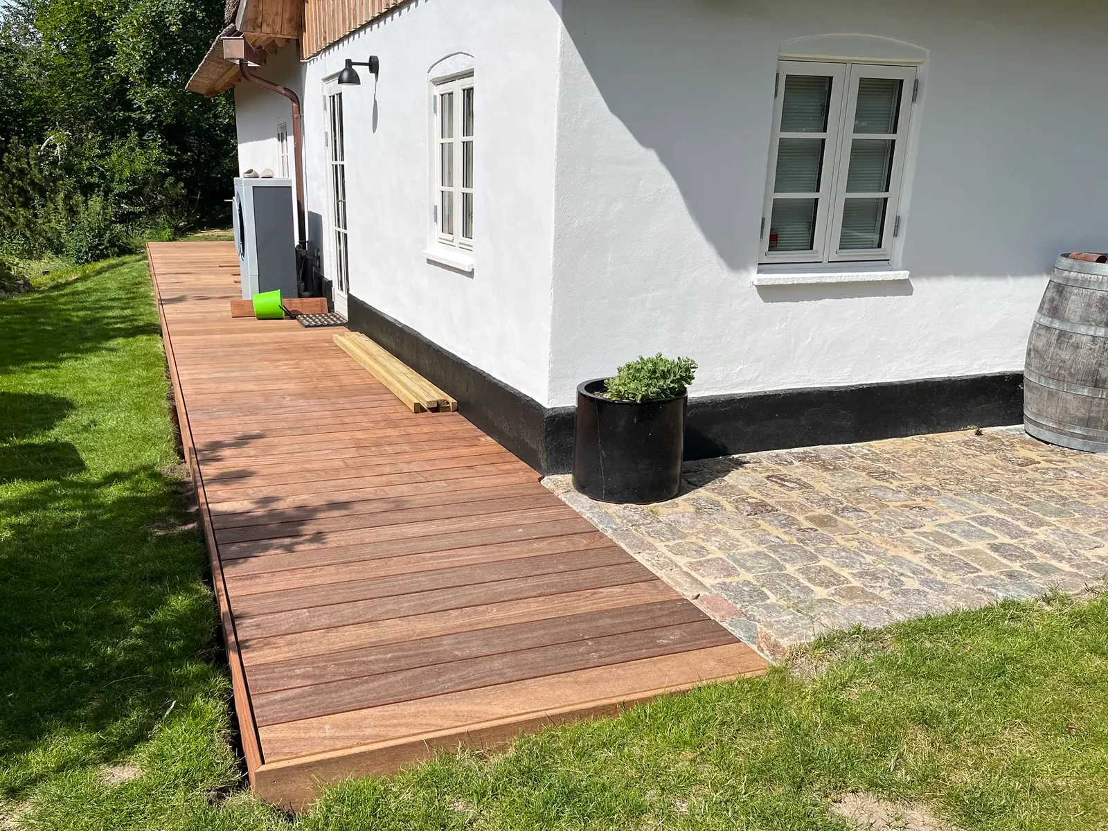 Terrasse i varmt, rødbrunt hårdttræ ført langs den hvide facade, hvor de nye brædder møder gammel brolægning ved husets hjørne