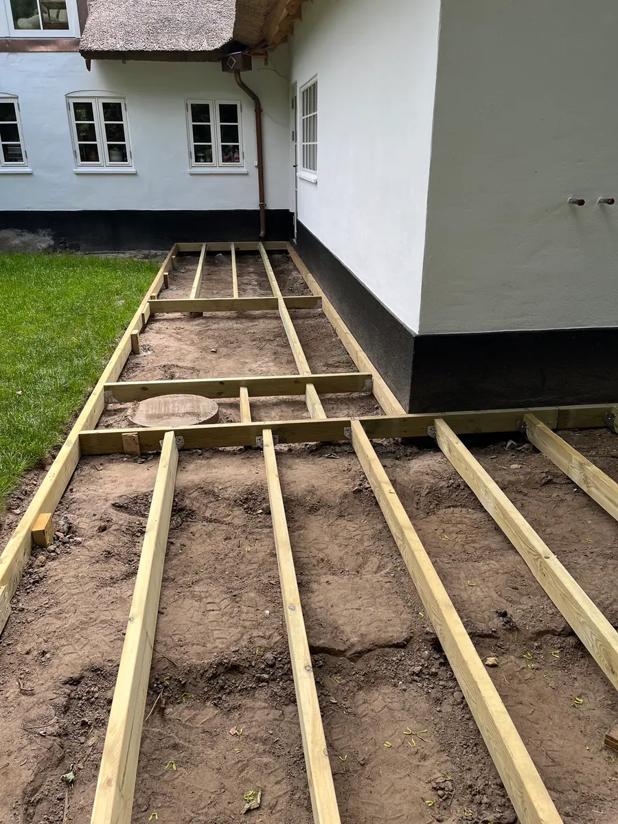 Strøer i lyst træ lagt ud i et rutenet på afrettet jord langs den hvide facade på et stråtækt hus, før terrassebrædderne kommer på